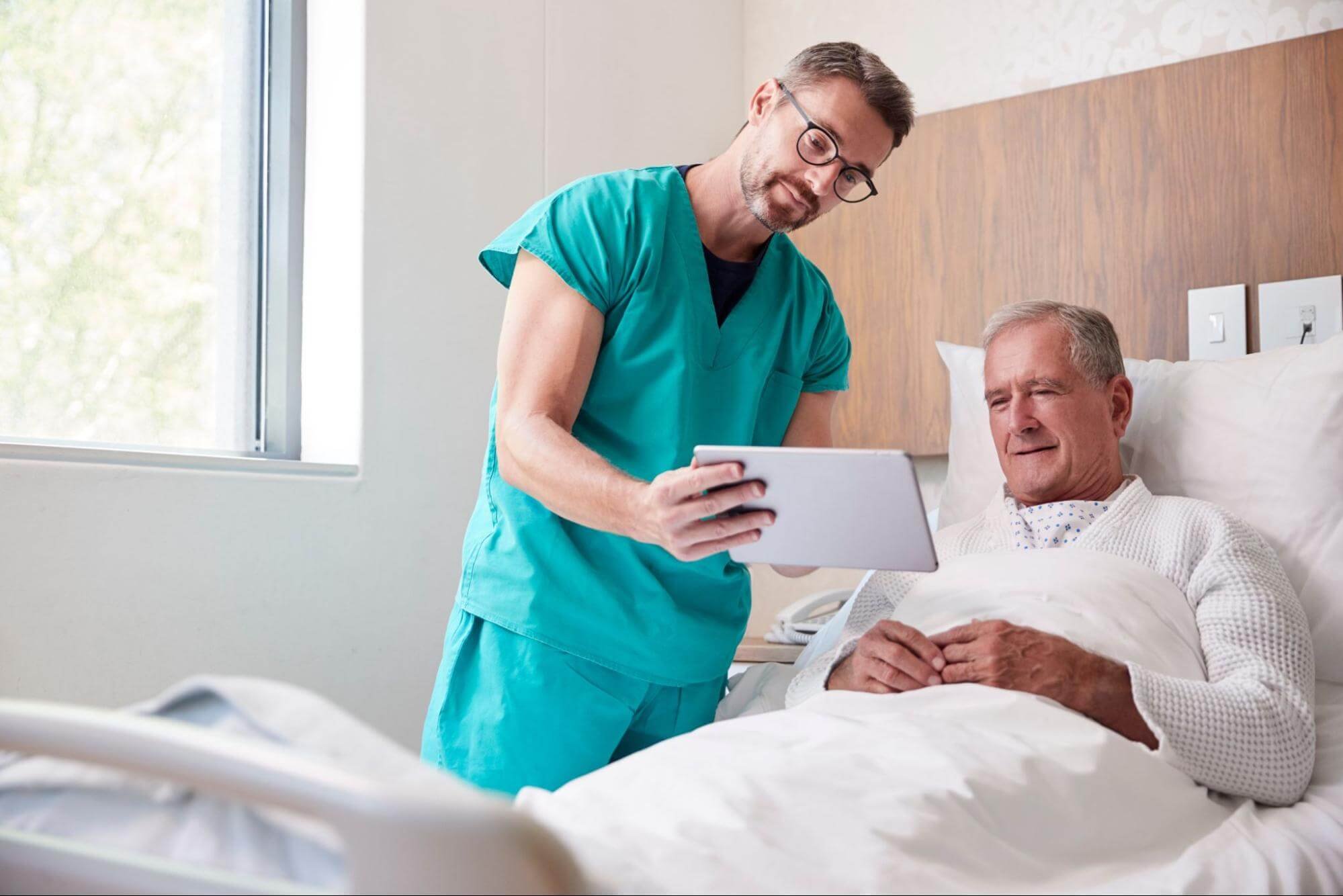 A physician wearing blue scrubs shows a tablet to an elderly patient who is in a hospital bed in front of wood cabinets
