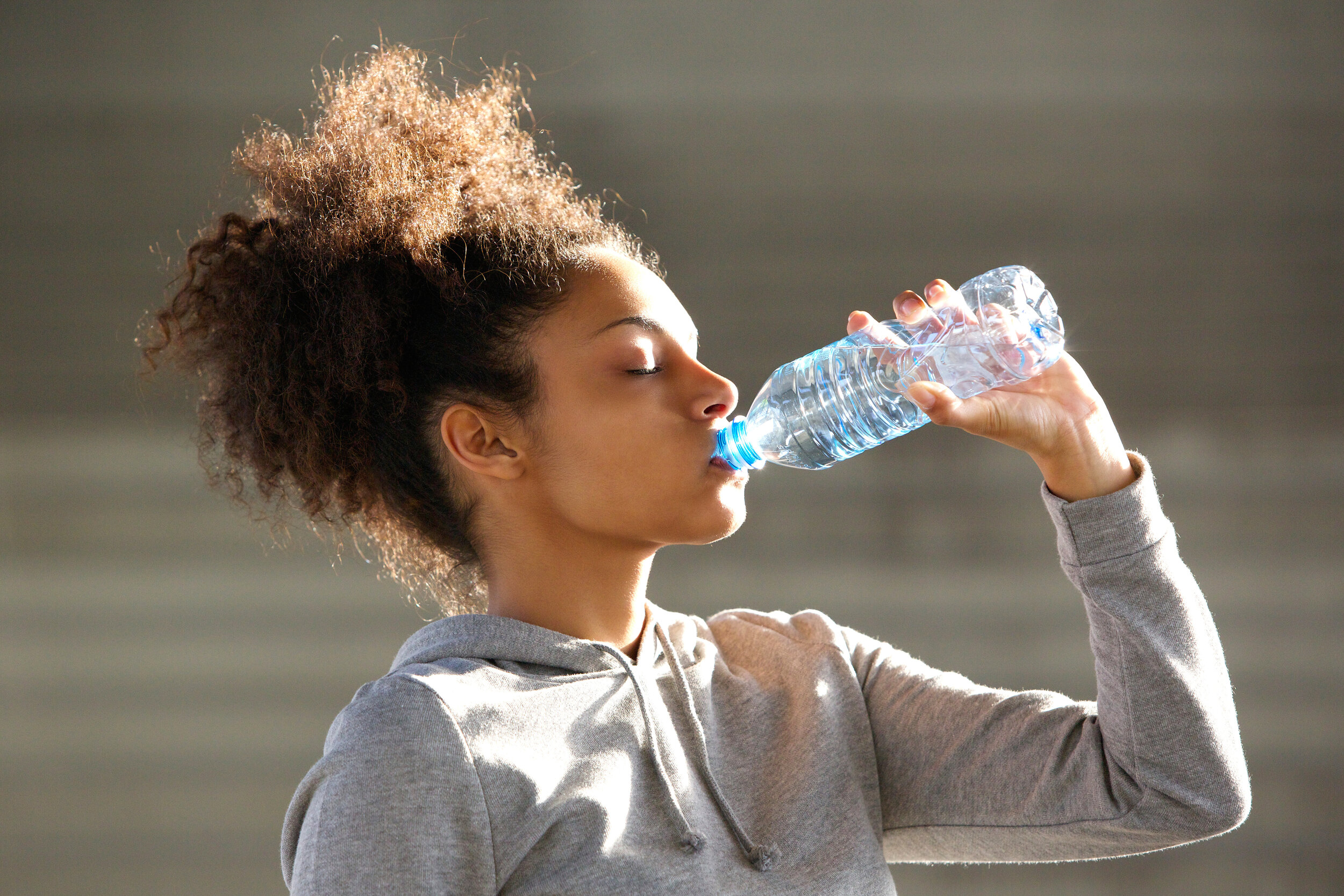 Young woman in gray hoodie drinking water in a plastic water bottle outdoors