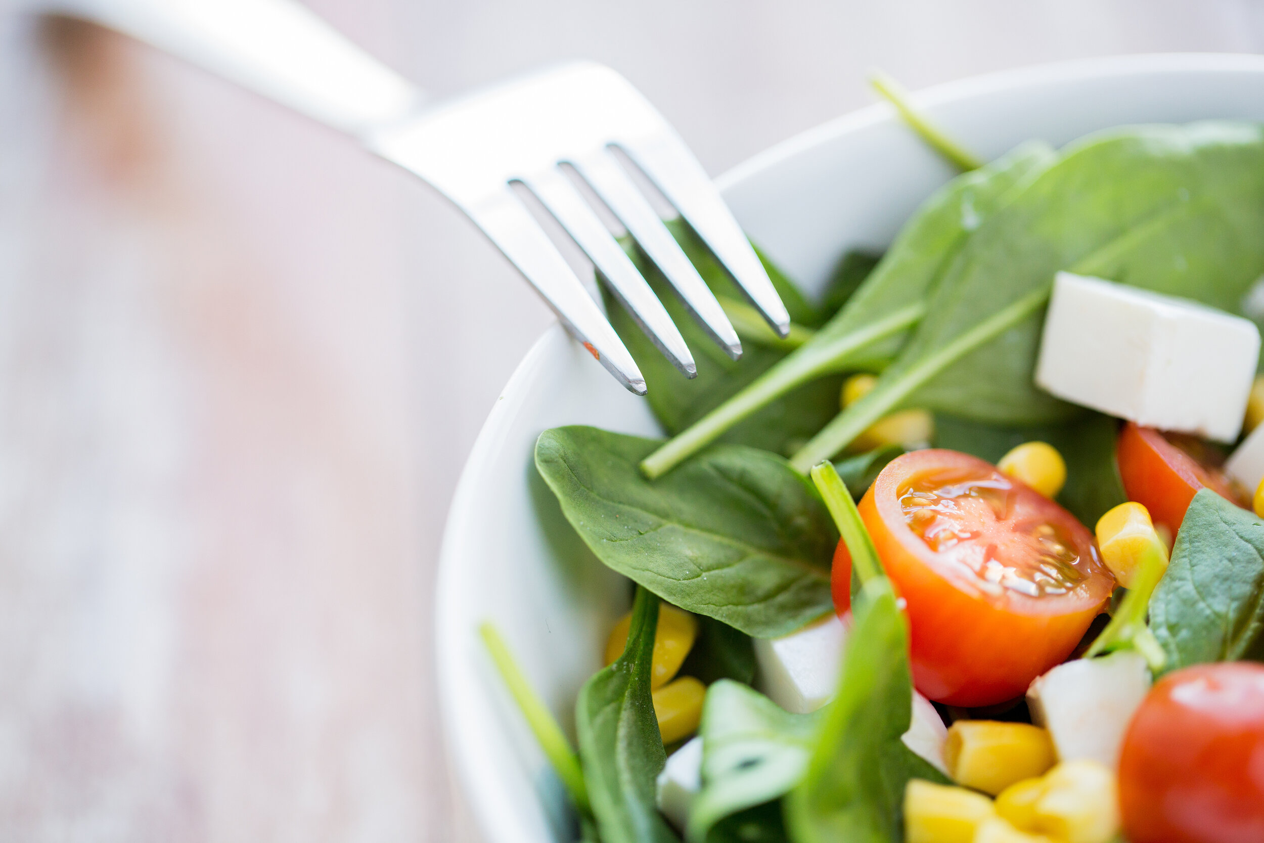 Close-up of spinach leaves, tomatoes, cheese, and corn as a salad in a white bowl with fork leaning on edge of bowl