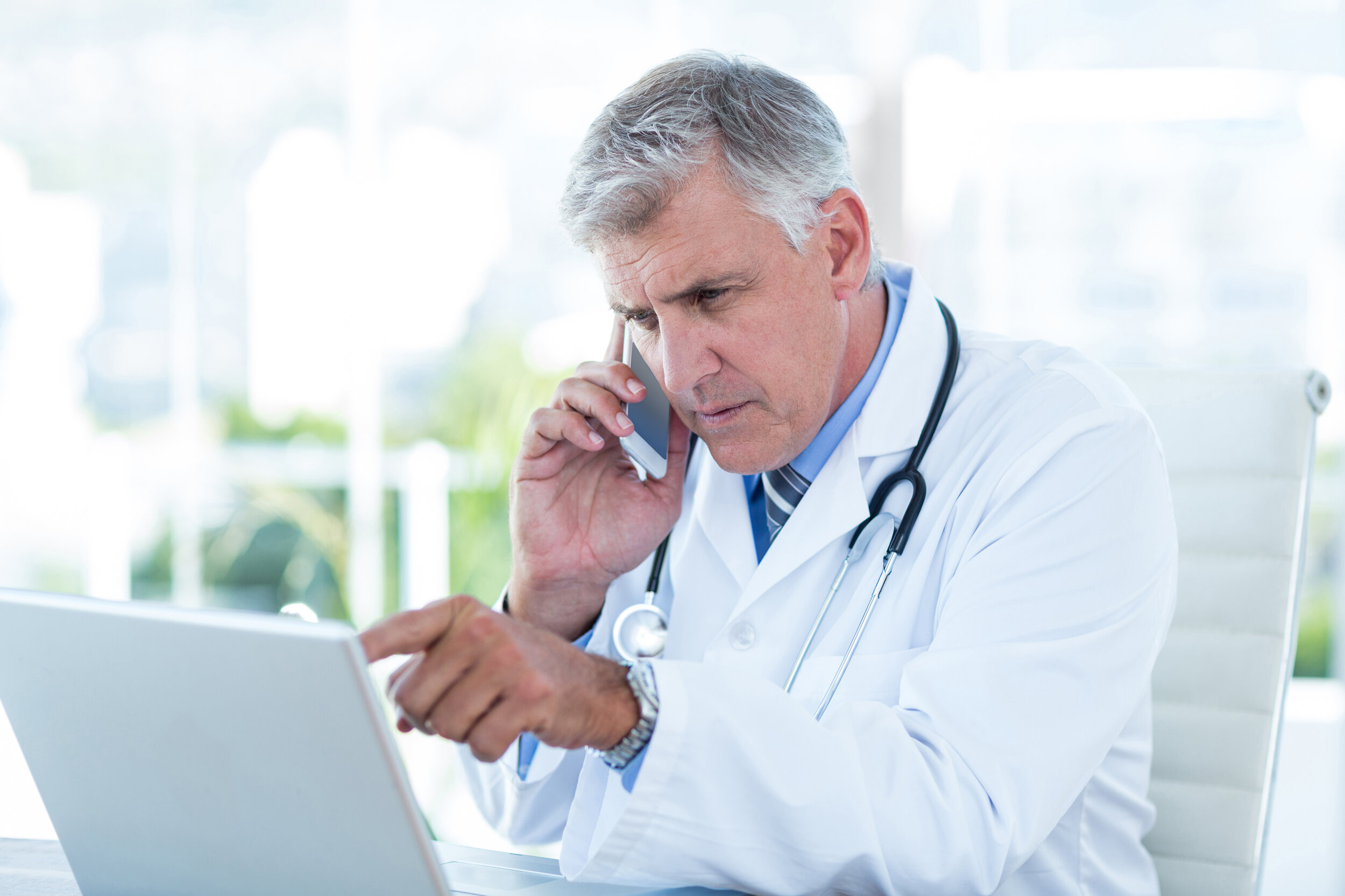 Serious male doctor working on laptop and talking to a patient over the phone.