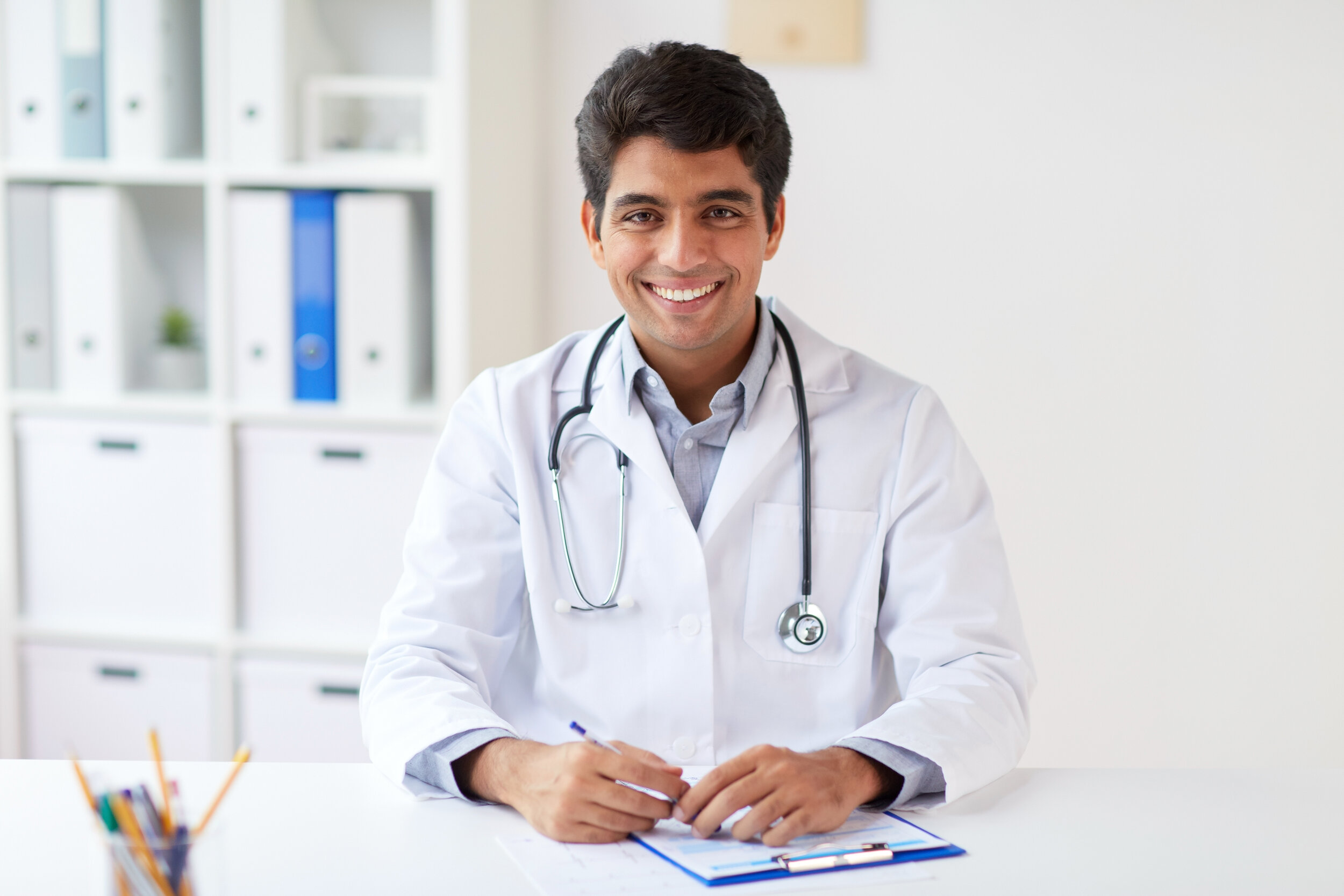 Smiling doctor in a white lab coat with stethoscope sitting at desk in a doctor’s office