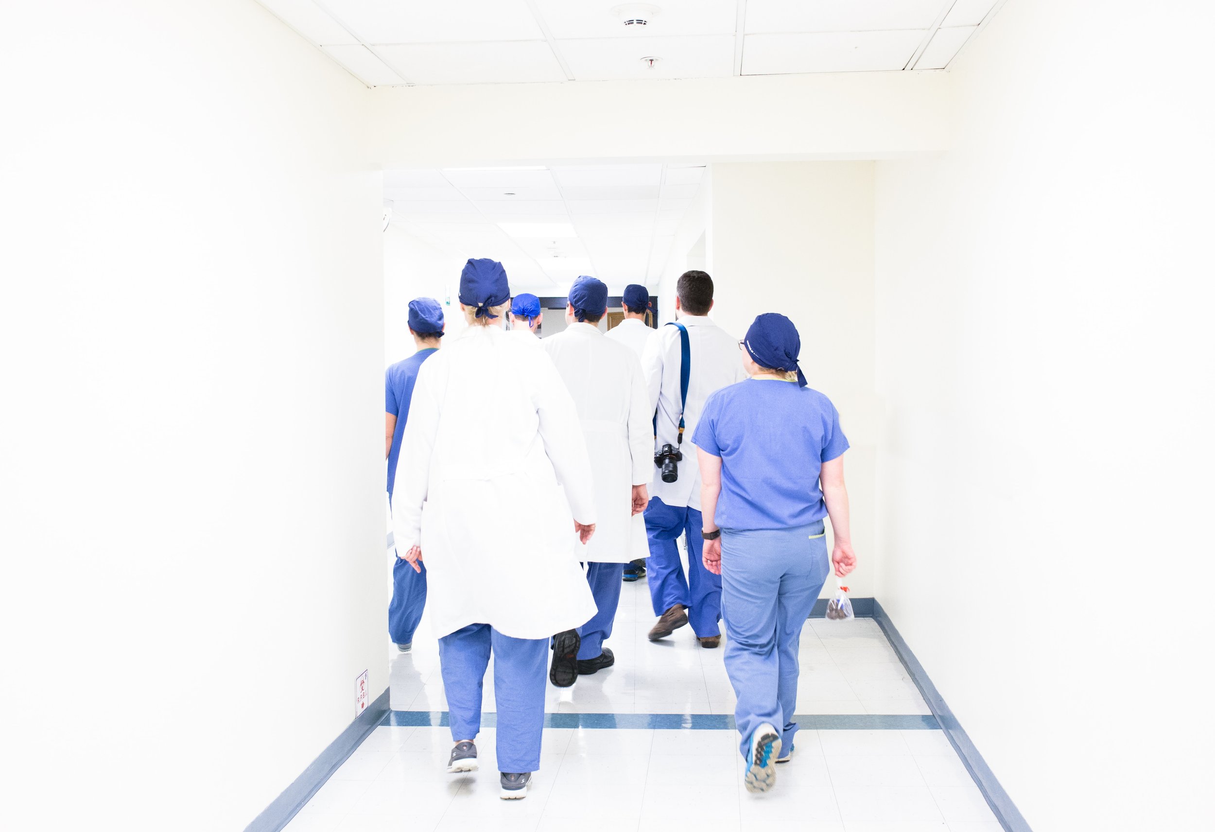 Doctors in white coats and scrub caps with nurses in blue scrubs walking down a white hall into an operating room